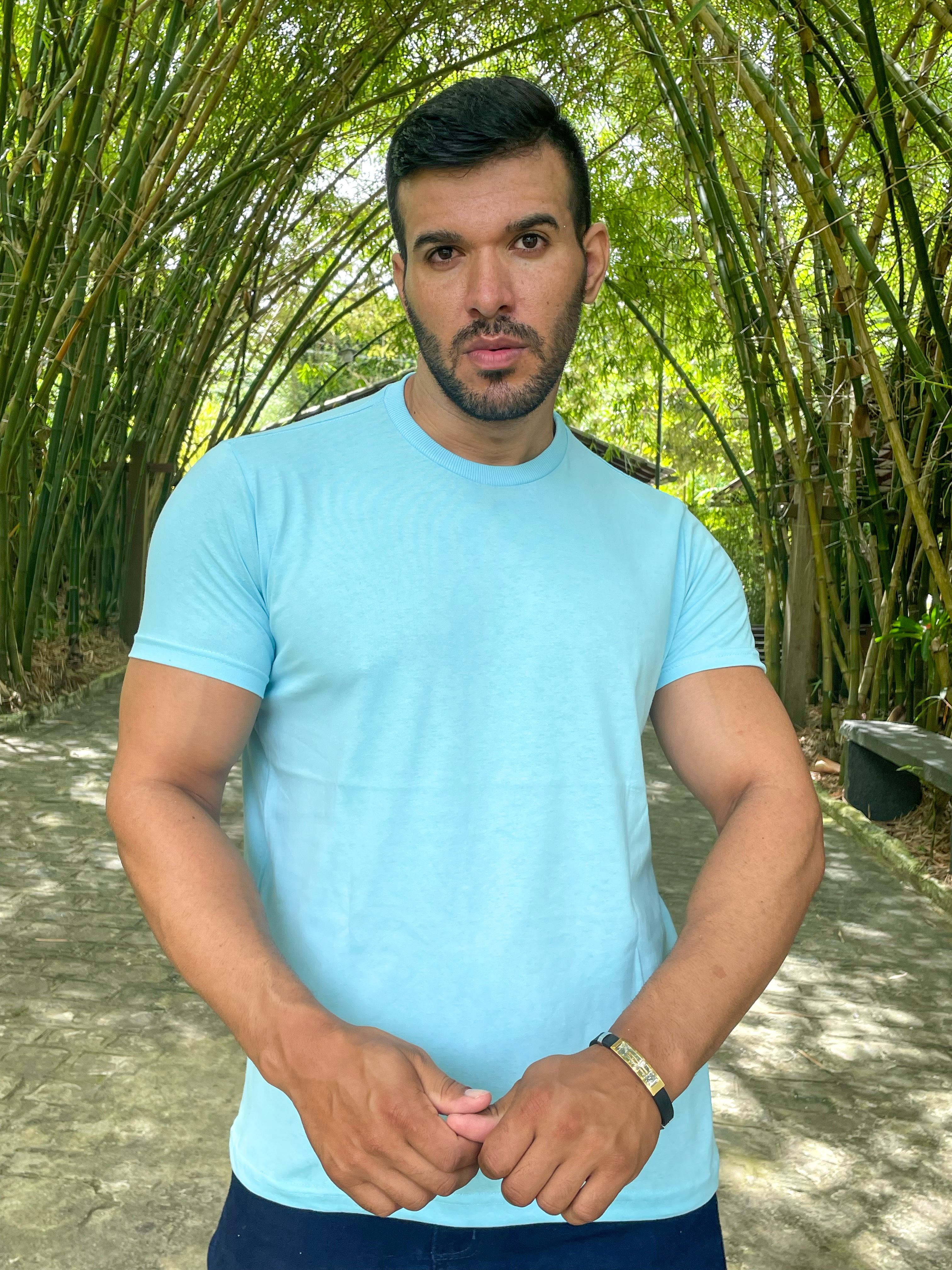 Homem posando em um ambiente natural, vestindo uma camiseta azul clara, com um fundo de bambus verdes.