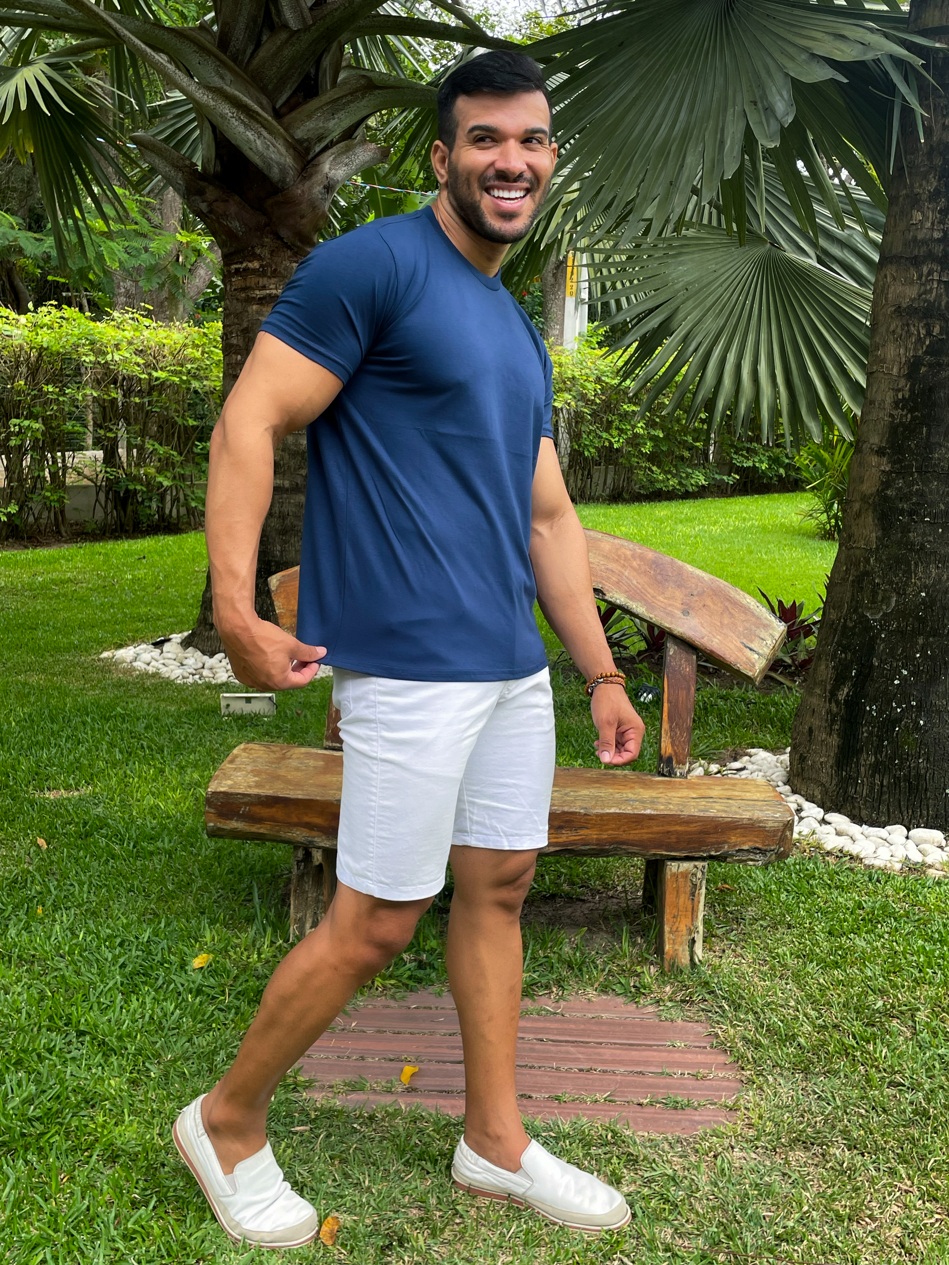 Homem sorridente com camiseta azul marinho e short branco, posando em um jardim tropical.