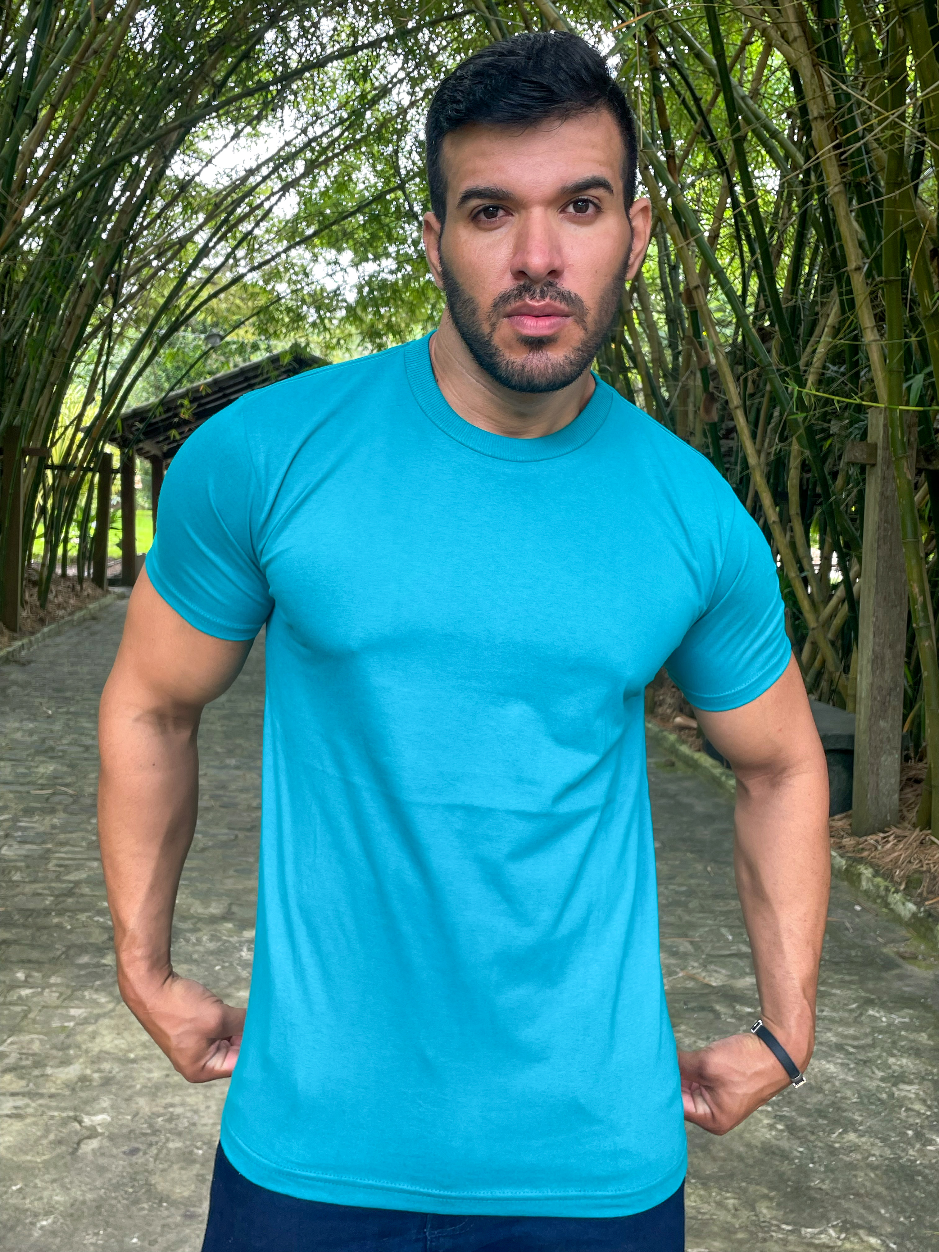 Homem posando em um ambiente natural, vestindo uma camiseta azul turquesa e com uma expressão intensa.