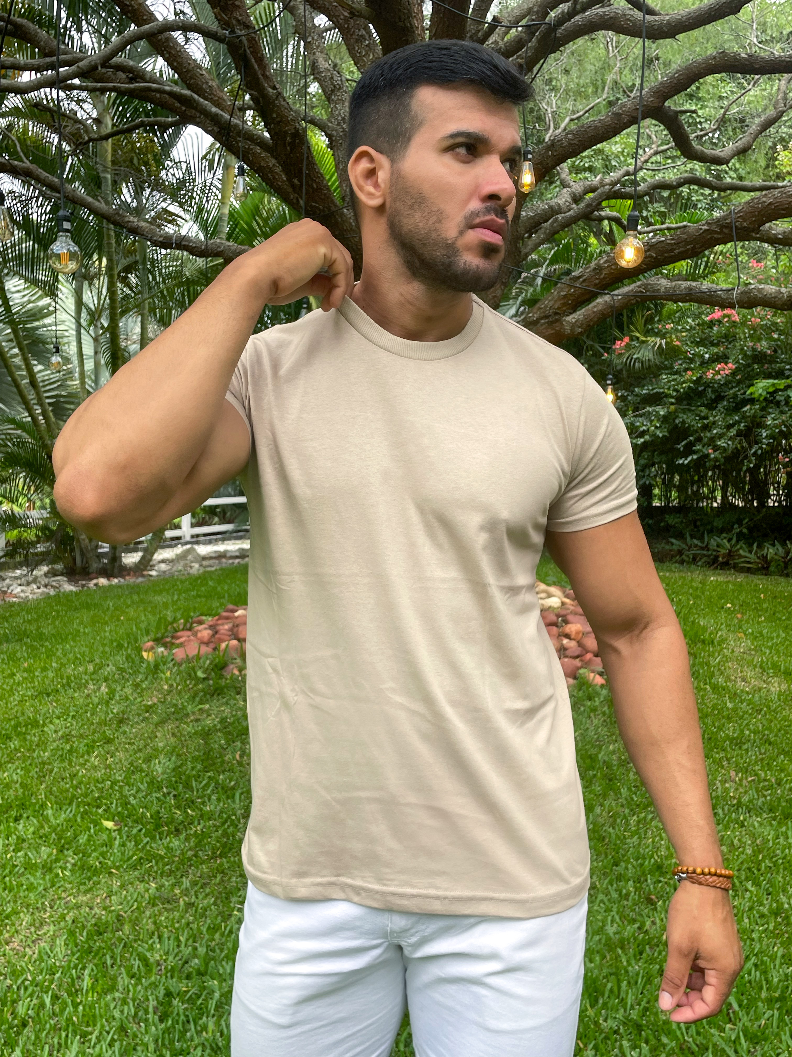 Homem posando ao ar livre, usando camiseta bege e bermuda branca, em um ambiente com árvores e luzes penduradas.