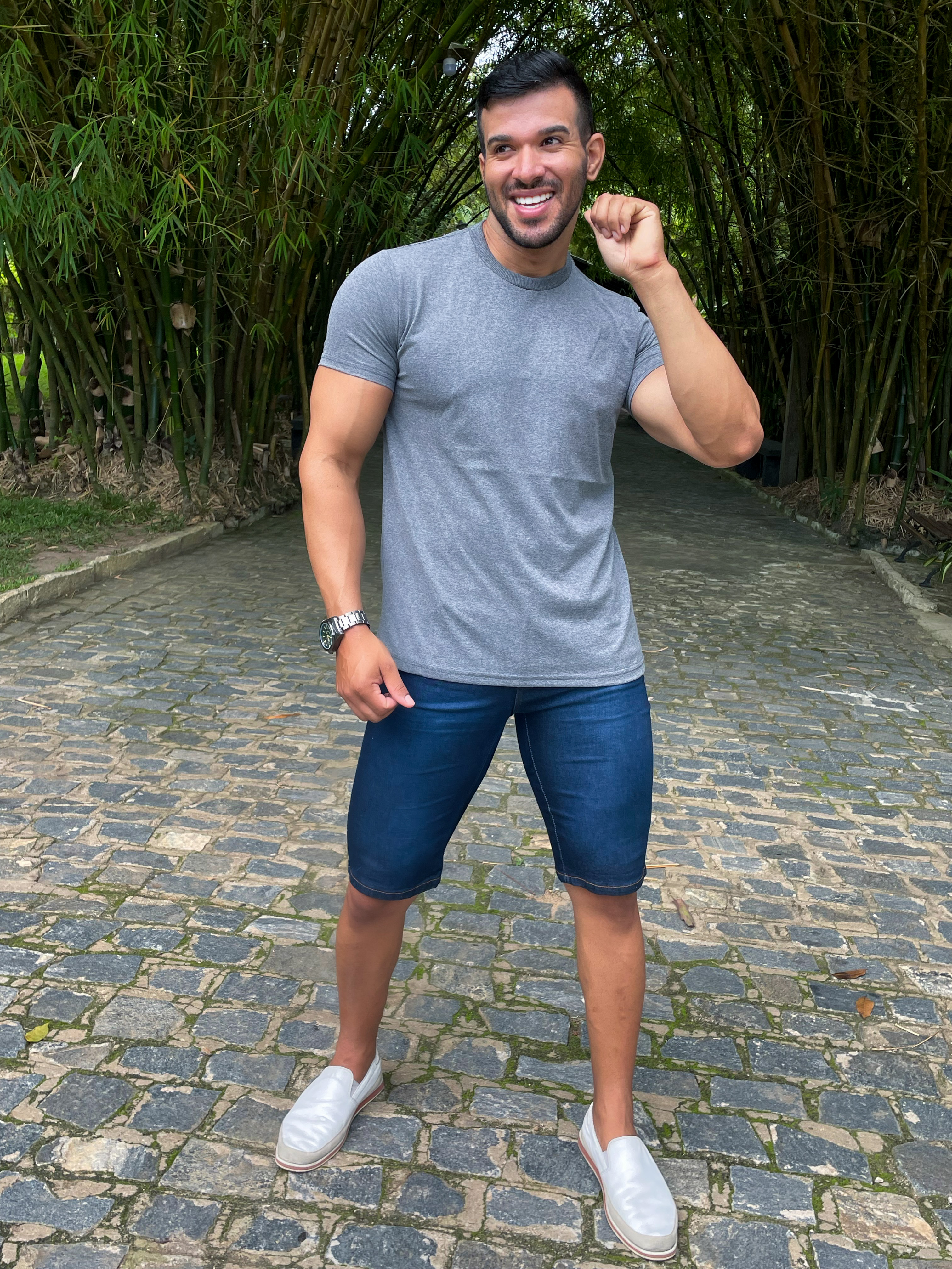 Homem sorridente caminhando em um caminho de pedras, vestindo uma camiseta cinza chumbo, bermuda jeans e sapatos brancos, rodeado por bambus verdes.