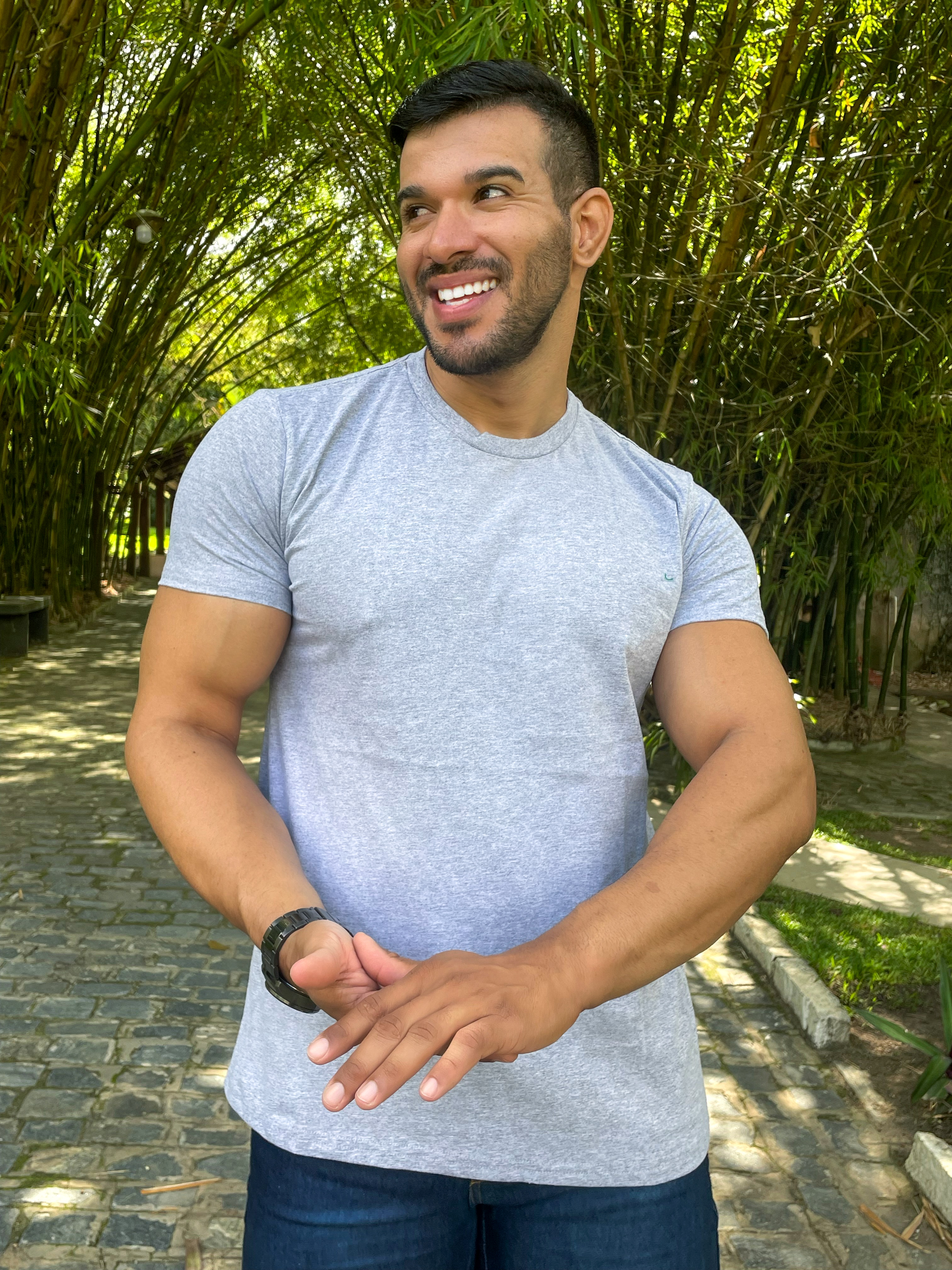 Homem sorridente em um ambiente natural, usando uma camiseta cinza mescla e jeans, posando ao ar livre entre bambus.