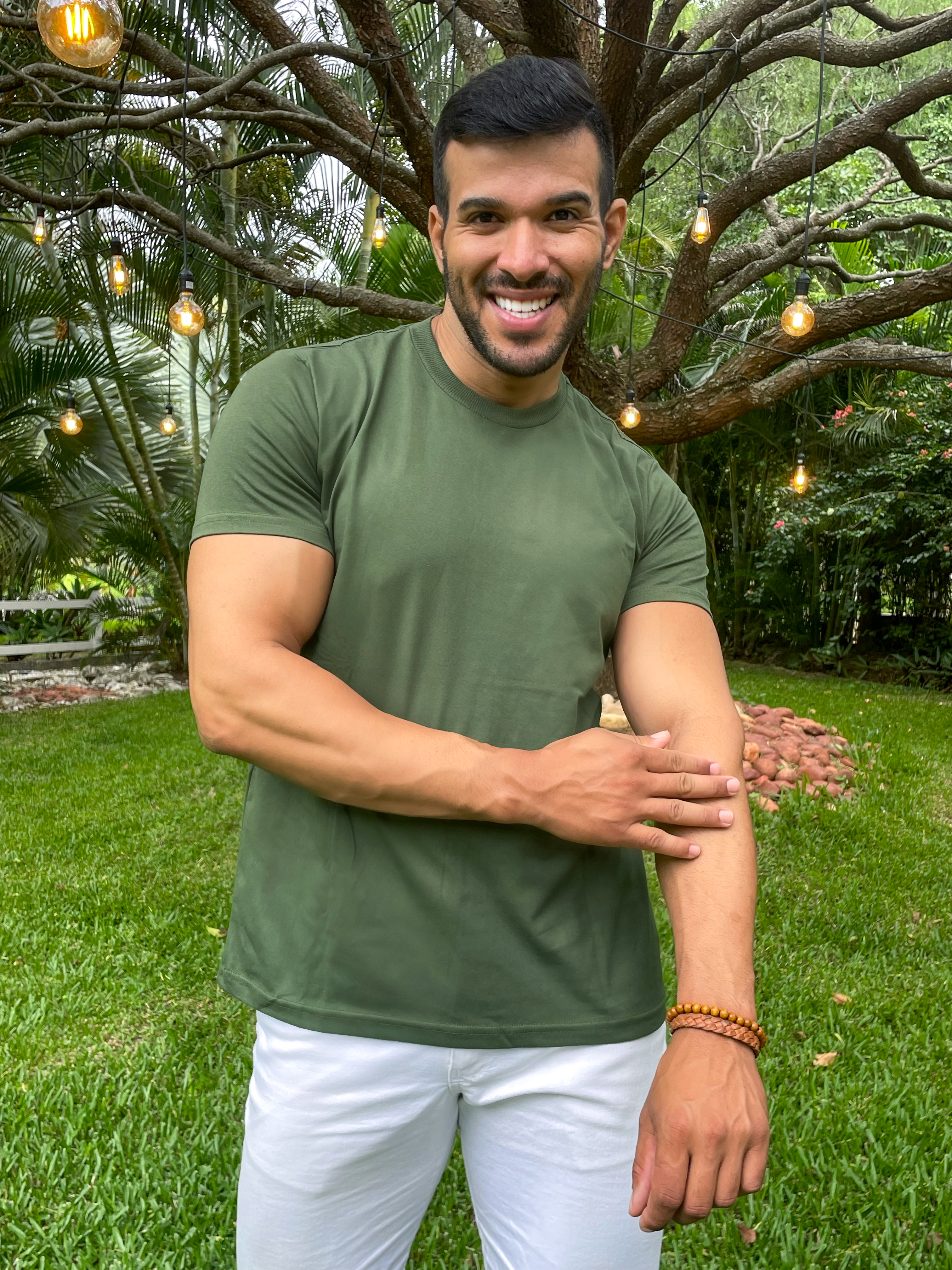 Homem sorridente vestindo camiseta verde em um ambiente ao ar livre, com árvores e luzes penduradas ao fundo.