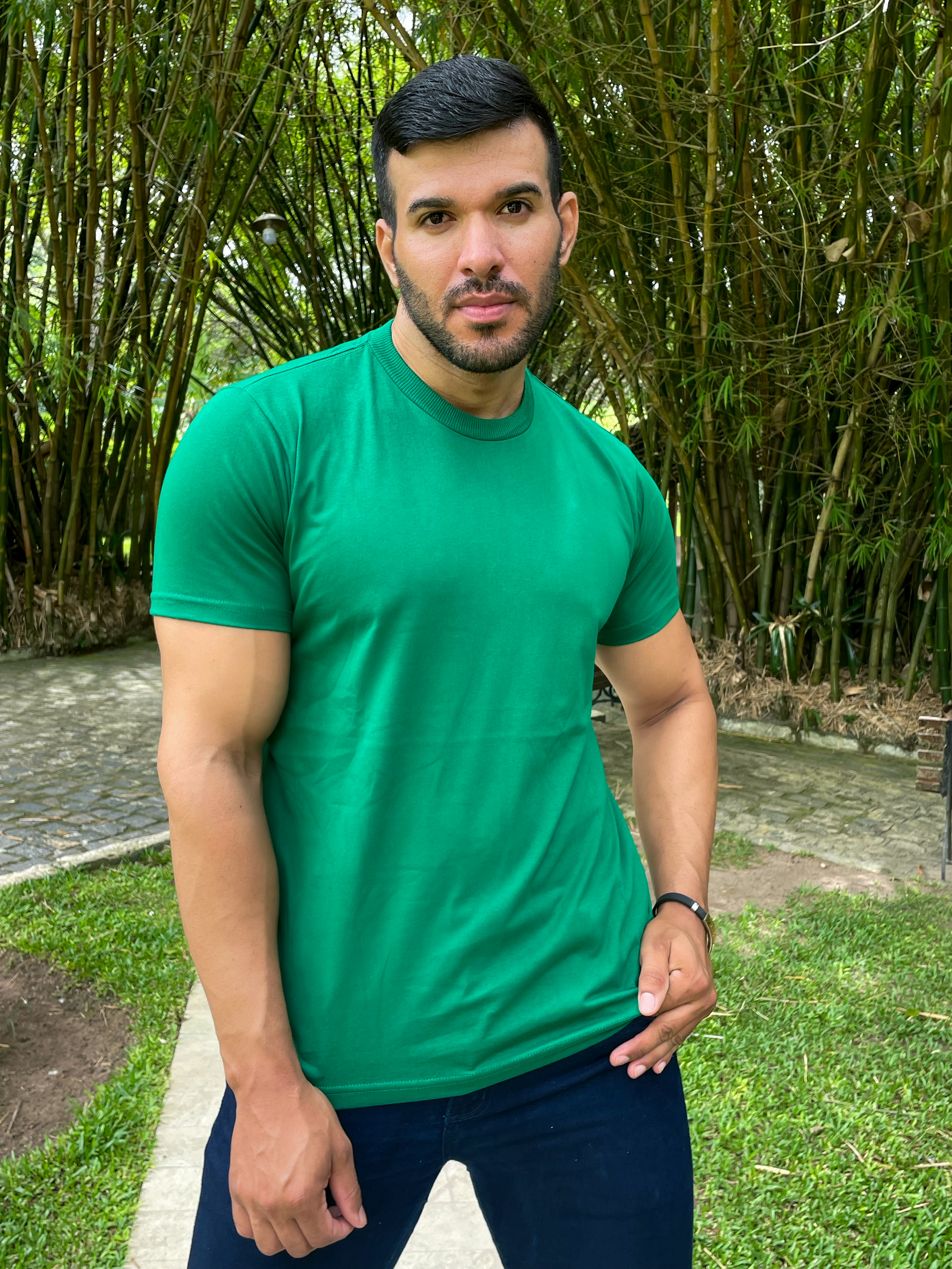 Homem posando em um ambiente externo, vestindo uma camiseta verde, com fundo de bambus e grama verde.