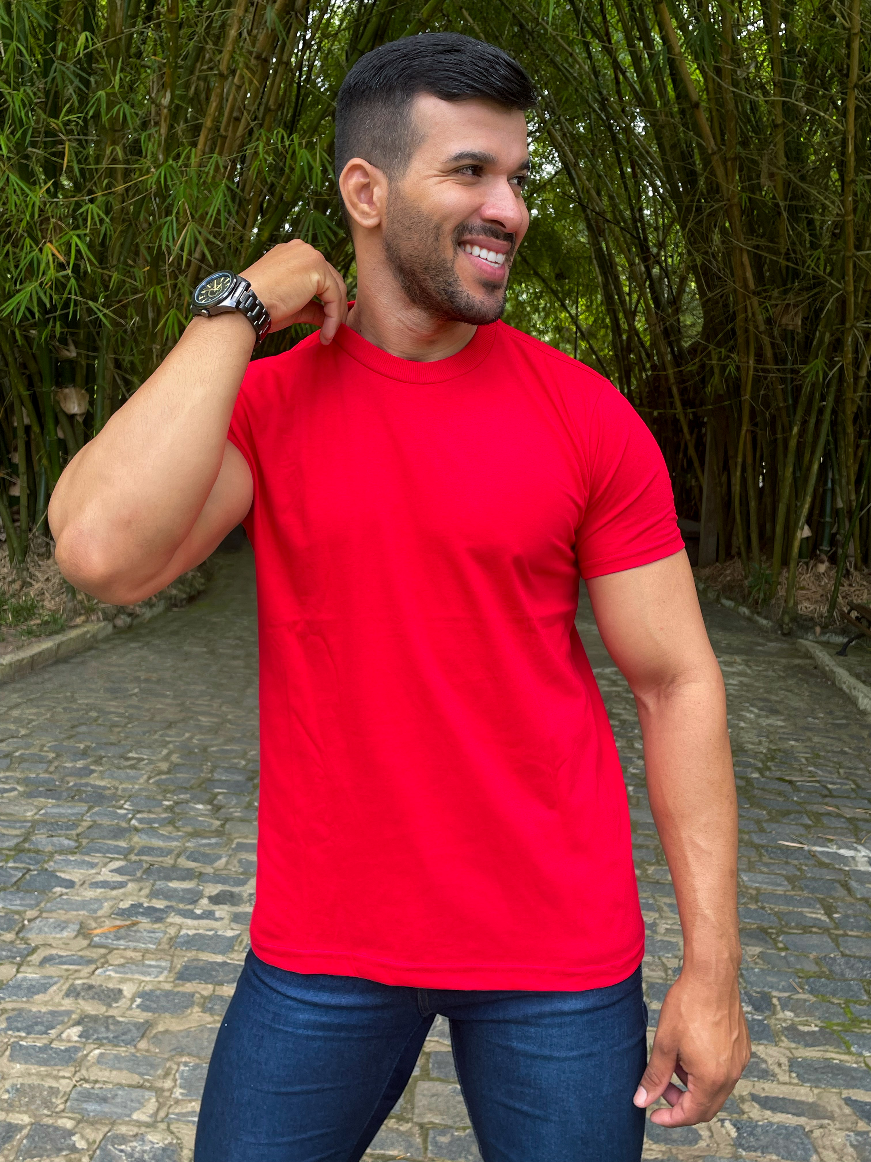 Homem sorrindo com camiseta vermelha em um ambiente natural com bambus ao fundo.