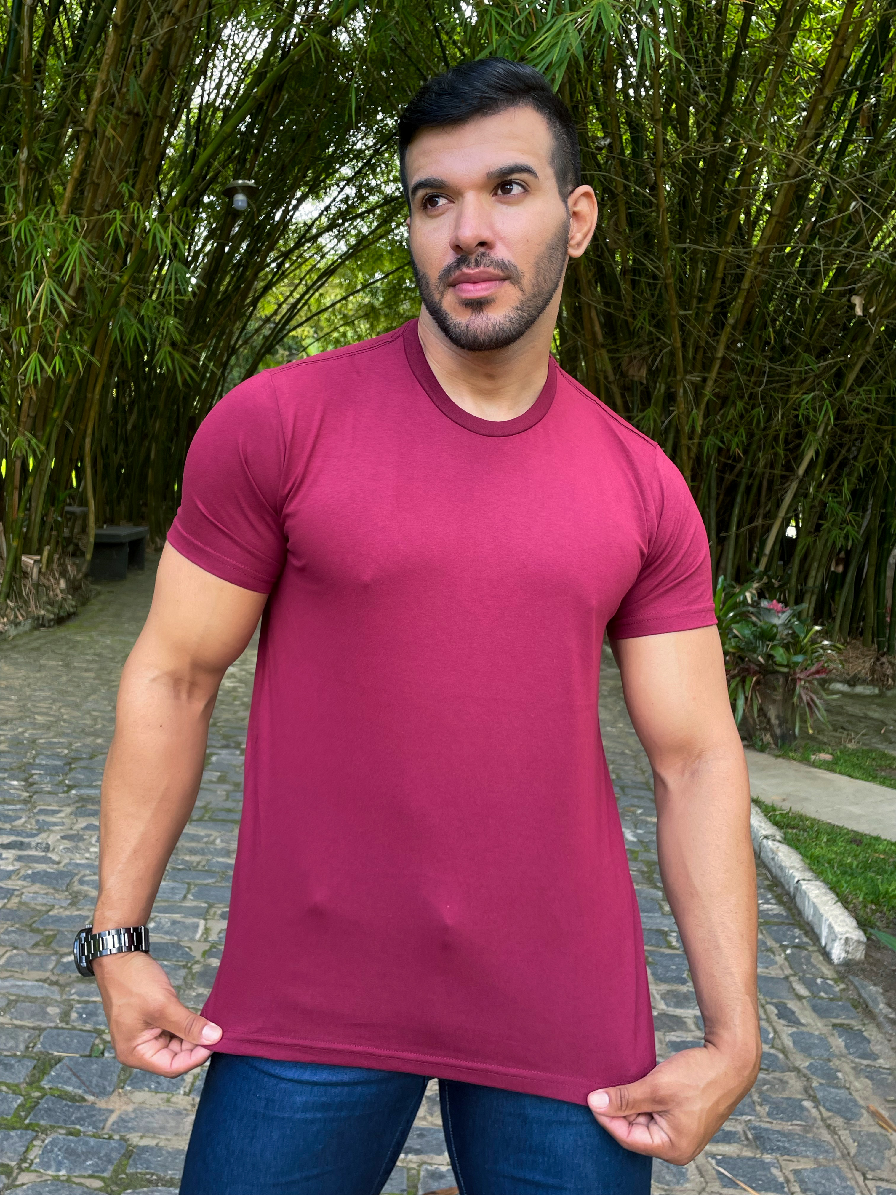 Homem posando ao ar livre, vestindo uma camiseta vinho e jeans, com um fundo de bambus verdes.